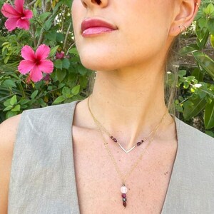 May include: A layered gold necklace with a V-shaped pendant and a pendant with a pink and red stone. The necklace is worn against a neutral-colored top, with pink flowers in the background.