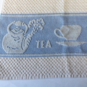 May include: Cream-colored kitchen towel with a textured weave. The towel features blue bands with embossed images of teacups, teapots, and the word "TEA" and "COFFEE".