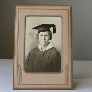 May include: A vintage black and white portrait photograph of a person wearing a graduation cap and gown with a white collar, encased in a rectangular tan cardboard frame featuring embossed geometric line patterns in the corners and borders.
