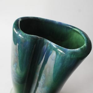 May include: A tall, ceramic vase with a glossy, flowing glaze in shades of green and blue. The vase has a unique, elongated oval opening and a gently curved body. The colors blend together, creating a marbled effect. The vase is a decorative item.