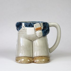 May include: A ceramic mug shaped like a person wearing overalls and holding a paintbrush. The mug is light gray with blue accents and brown shoes.