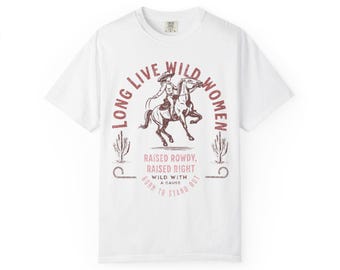 May include: A white short-sleeve t-shirt featuring a maroon graphic of a person riding a horse. The text reads LONG LIVE WILD WOMEN, RAISED ROWDY, RAISED RIGHT, WILD WITH A CAUSE, and BORN TO STAND OUT, flanked by two cactus illustrations.