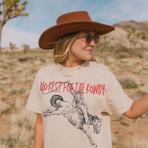 May include: A beige t-shirt with a black and white illustration of a cowboy riding a bucking horse. The text "NO REST FOR THE ROWDY" is printed in red on the shirt.