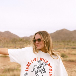 May include: A white t-shirt with a graphic of a woman riding a horse. The graphic is surrounded by the text 'Long Live Wild Women' in a red and black circular design. The text below the graphic reads 'Raised Rowdy, Raised Right, Wild with a Cause, Born to Stand Out.' Long Live Wild Women | Womens Rights Vintage Comfort Colors Shirt, Western Grunge Cowgirl, Resist Feminist Graphic Crewneck, Freedom Shirt