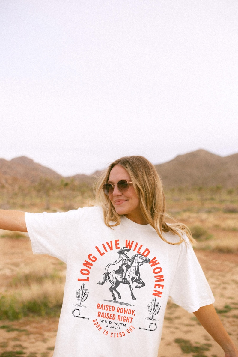 May include: A white t-shirt with a graphic of a woman riding a horse. The graphic is surrounded by the text 'Long Live Wild Women' in a red and black circular design. The text below the graphic reads 'Raised Rowdy, Raised Right, Wild with a Cause, Born to Stand Out.' Long Live Wild Women | Womens Rights Vintage Comfort Colors Shirt, Western Grunge Cowgirl, Resist Feminist Graphic Crewneck, Freedom Shirt