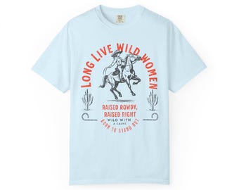 May include: A light blue t-shirt featuring a graphic print of a person riding a horse. The text reads LONG LIVE WILD WOMEN, RAISED ROWDY, RAISED RIGHT, WILD WITH A CAUSE, and BORN TO STAND OUT. The design includes illustrations of two cacti.