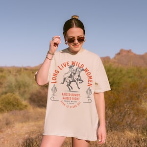 A beige t-shirt with the text 'Long Live Wild Women' in a red and black font. The text is surrounded by a circle.  Black and white illustration of a woman riding a horse. Below the circle is the text 'Raised Rowdy, Raised Right' in red font. Below that is the text 'Wild With A Cause' in black font. Below that is the text 'Born To Stand Out' in red font. Long Live Wild Women | Womens Rights Vintage Comfort Colors Shirt, Western Grunge Cowgirl, Resist Feminist Graphic Crewneck, Freedom Shirt