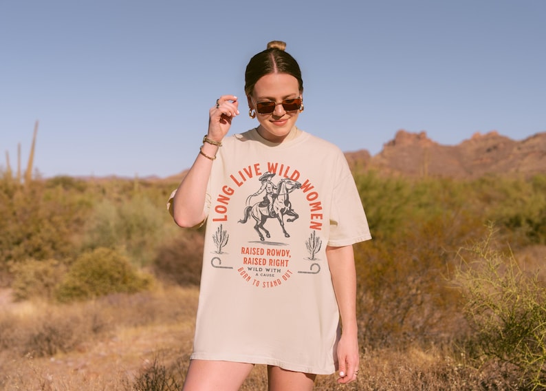A beige t-shirt with the text 'Long Live Wild Women' in a red and black font. The text is surrounded by a circle.  Black and white illustration of a woman riding a horse. Below the circle is the text 'Raised Rowdy, Raised Right' in red font. Below that is the text 'Wild With A Cause' in black font. Below that is the text 'Born To Stand Out' in red font. Long Live Wild Women | Womens Rights Vintage Comfort Colors Shirt, Western Grunge Cowgirl, Resist Feminist Graphic Crewneck, Freedom Shirt