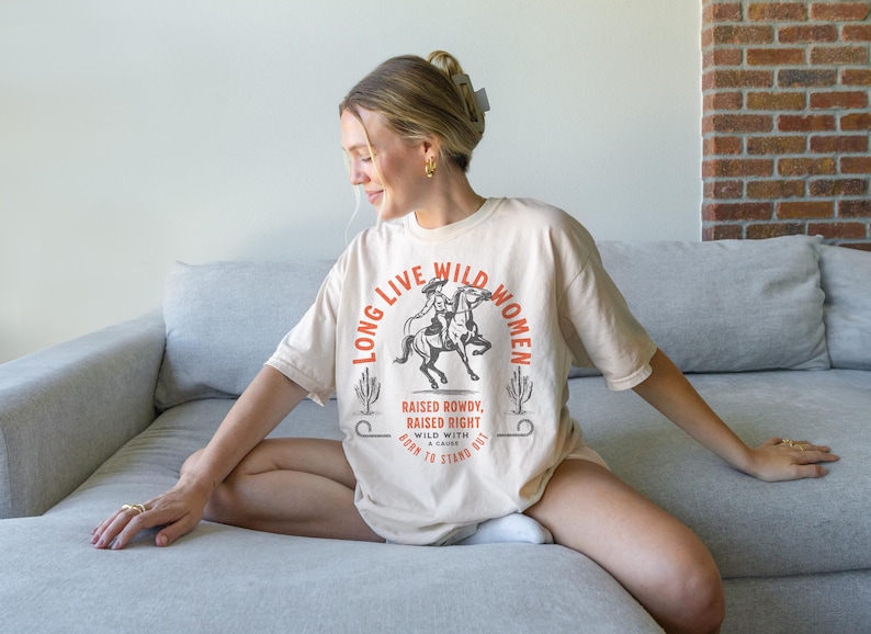 May include: A beige t-shirt with the text 'Long Live Wild Women' in orange. The text is surrounded by a graphic of a woman riding a horse. The text below the graphic reads 'Raised Rowdy, Raised Right. Wild with a Cause. Born to Stand Out.' Long Live Wild Women | Womens Rights Vintage Comfort Colors Shirt, Western Grunge Cowgirl, Resist Feminist Graphic Crewneck, Freedom Shirt