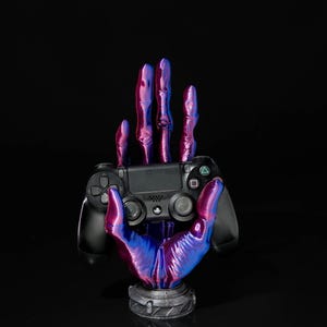 May include: A black PlayStation 4 controller held by a stylized hand-shaped stand. The stand is painted with a gradient of blue and purple hues. The base of the stand is a dark gray color, and the background is black.
