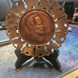 May include: A gold-colored metal plaque with a bronze-colored portrait of a man with a beard and the text "Tchaikovsky" on the bottom. The plaque is surrounded by a decorative metal frame with butterfly designs and is on a black stand.