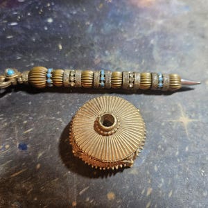 May include: A gold-toned pen with blue and white accents and a matching decorative stand. The pen has a decorative design with small beads and a pointed tip. The stand has a circular base with a raised center.