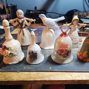 May include: Seven porcelain bells with various designs.  The bells feature a blue bird, a girl in a white dress, a girl in a red dress, a girl in a brown dress, a floral design, a floral design with the text "Tennessee", and a floral design with the text "South Carolina".