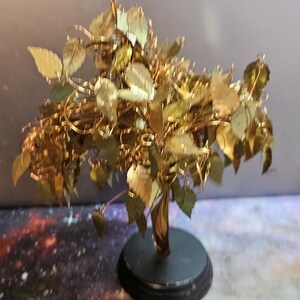 May include: A gold-colored metal tree sculpture with a twisted trunk and many leaves. The tree is on a black circular base.