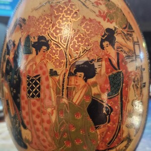 May include: A ceramic egg-shaped trinket box with a painted scene of four women in traditional Japanese clothing standing in front of a flowering tree. The background is a light brown with gold accents.