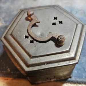 May include: A vintage, hexagonal metal box with a hinged lid and a decorative handle. The box is a muted gray color with a darker, aged handle. The lid features small, cross-shaped cutouts. The box appears to be antique.