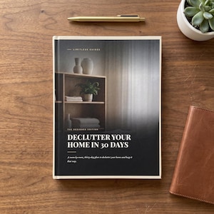 Może przedstawiać: Książka w twardej oprawie zatytułowana Declutter Your Home in 30 Days z okładką przedstawiającą drewnianą półkę z wazonem, rośliną doniczkową i złożonymi ręcznikami. Książka posiada ciemną dolną część z białym tekstem.