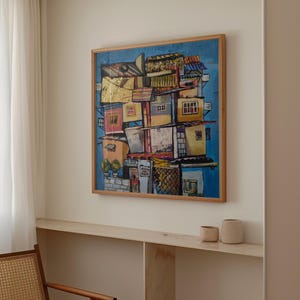 May include: A framed abstract painting depicting a colorful cityscape with stacked buildings. The artwork features a blue background and a wooden frame. Gold leaf accents add a touch of luxury to the piece. The painting is displayed on a white wall.