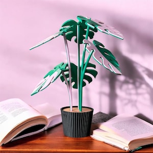 May include: A decorative plant sculpture featuring dark green, layered monstera leaves with white edges, supported by thin green and white stems. The plant is set in a black, vertically ribbed, cylindrical pot with a flat, light-colored top surface.