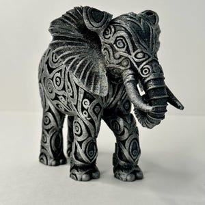 May include: A detailed silver elephant figurine with intricate patterns. The elephant has large ears, a long trunk, and tusks. The body is covered in swirling, circular designs, giving it a textured appearance. The figurine is standing on a white surface.