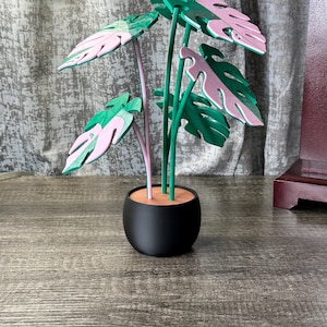 May include: A decorative artificial plant in a black pot. The plant features green and pink leaves with green stems. The pot is round and sits on a wooden surface. The leaves have a split leaf design.