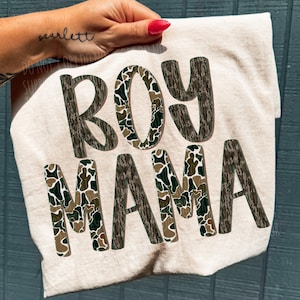 May include: A light beige t-shirt with the words "BOY MAMA" printed in a camouflage pattern. The letters are a mix of brown, green, and tan. The shirt is held up by a hand with a tattoo and red nail polish.