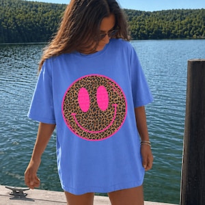 May include: A periwinkle blue short-sleeve t-shirt featuring a large circular graphic on the front. The graphic consists of a leopard print smiley face with two oval eyes and a curved mouth, all outlined in a vibrant neon pink border.