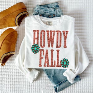 May include: A white long-sleeve crewneck sweatshirt featuring the text HOWDY FALL in a reddish-brown paisley pattern. Two turquoise-colored floral concho graphics are positioned next to the text. The sweatshirt is paired with blue denim jeans.