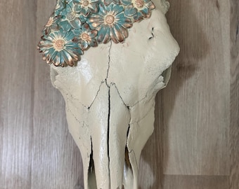May include: A white animal skull with a floral design. The flowers are made of a light blue and gold material and are arranged in a crown shape on top of the skull.