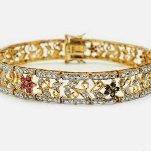 May include: A gold link bracelet featuring an intricate filigree design with embedded clear stones. The piece includes floral motifs accented with small red, blue, and green gemstones set within the metalwork, finished with a secure box clasp.