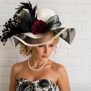 May include: A cream-coloured hat with a wide brim featuring a black and white pattern. The hat is decorated with black feathers, a black sheer bow, and a deep red flower. A pearl necklace is also visible.