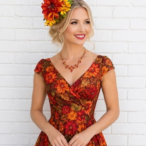Op de afbeelding: Een jurk met bloemenprint in een levendig patroon van rode, oranje en gele bloemen met groene bladeren. De jurk heeft een V-hals en kapmouwtjes. Een bijpassend hoofddeksel met kunstbloemen en een zwart spiraalvormig accent wordt gedragen.