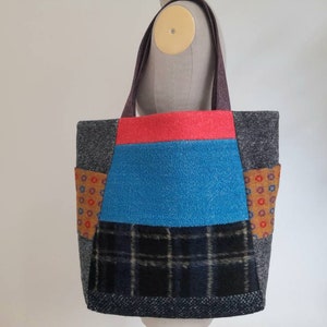 May include: A colourful patchwork tote bag. The bag is made from various fabrics, including red, blue, and black plaid. It has grey and orange accents, and the handles are a dark purple colour.