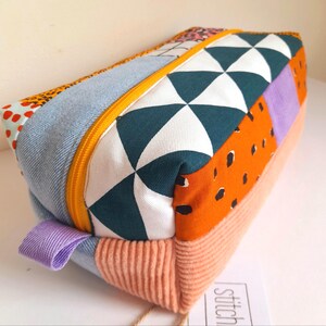 May include: A rectangular patchwork pencil case with a yellow zip and a lavender pull tab. The case features various fabric patterns, including denim, geometric shapes, and orange with black spots. The bottom section is peach corduroy.