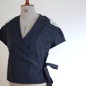May include: A dark blue denim wrap top featuring short sleeves and a V-neckline. The shoulders are accented with a white fabric panel featuring a small black floral pattern. The garment includes a side tie closure at the waist for a secure fit.