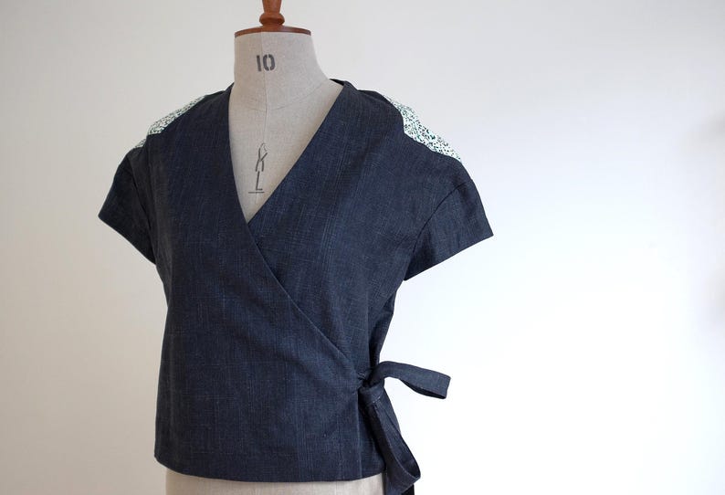 May include: A dark blue denim wrap top featuring short sleeves and a V-neckline. The shoulders are accented with a white fabric panel featuring a small black floral pattern. The garment includes a side tie closure at the waist for a secure fit.