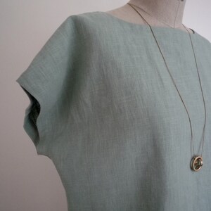 May include: A light blue linen top with a boat neck and short sleeves. A long, thin gold-colored necklace with a circular pendant hangs on the top. The top is displayed on a mannequin.