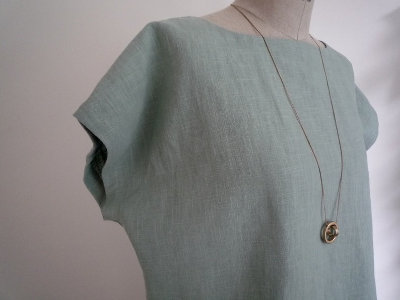 May include: A light blue linen top with a boat neck and short sleeves. A long, thin gold-colored necklace with a circular pendant hangs on the top. The top is displayed on a mannequin.