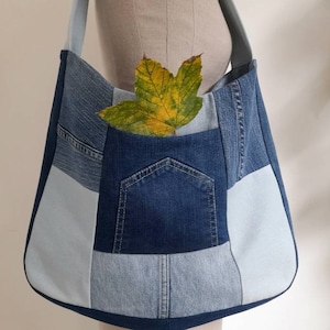 May include: A large blue denim tote bag with a patchwork design. The bag has a large pocket on the front and a light blue fabric lining. A single yellow leaf is placed inside the bag.