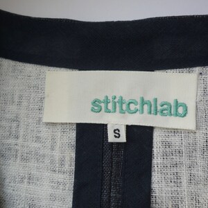 May include: A garment featuring a white fabric label with the teal embroidered text stitchlab and a smaller white tag below it displaying the letter S in black. The garment has a dark navy blue trim along the neckline and a textured white fabric.
