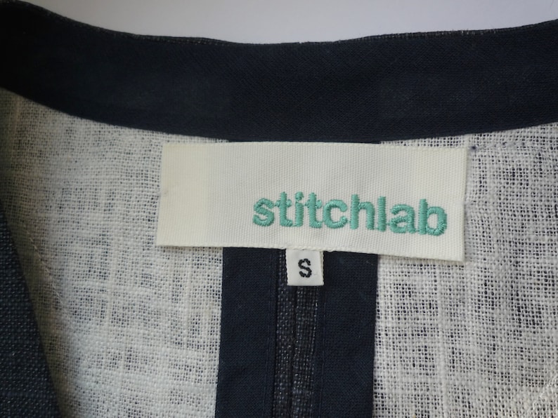 May include: A garment featuring a white fabric label with the teal embroidered text stitchlab and a smaller white tag below it displaying the letter S in black. The garment has a dark navy blue trim along the neckline and a textured white fabric.