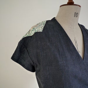 May include: A dark charcoal gray wrap top with short sleeves and a V-neckline. The shoulders feature contrasting white panels with a speckled pattern of small green and black dots. The fabric has a visible woven texture throughout the garment.