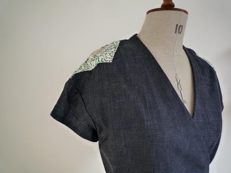 May include: A dark charcoal gray wrap top with short sleeves and a V-neckline. The shoulders feature contrasting white panels with a speckled pattern of small green and black dots. The fabric has a visible woven texture throughout the garment.