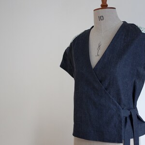 May include: A dark blue wrap top with short sleeves and a V-neckline. The garment features a textured fabric finish and light-colored patterned fabric panels on the shoulders. It includes a side tie closure at the waist for a secure fit.