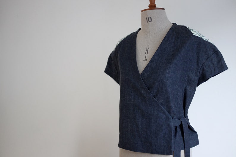 May include: A dark blue wrap top with short sleeves and a V-neckline. The garment features a textured fabric finish and light-colored patterned fabric panels on the shoulders. It includes a side tie closure at the waist for a secure fit.