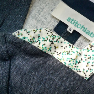 May include: A dark charcoal gray garment with a textured weave featuring a white collar accent decorated with a small green and black speckled pattern. A white fabric label is stitched at the neckline with the text stitchlab and a size S tag.
