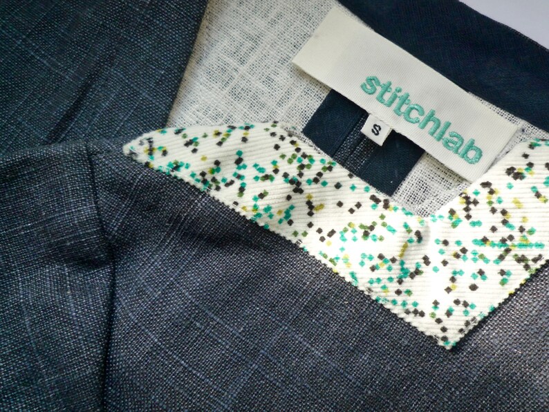 May include: A dark charcoal gray garment with a textured weave featuring a white collar accent decorated with a small green and black speckled pattern. A white fabric label is stitched at the neckline with the text stitchlab and a size S tag.