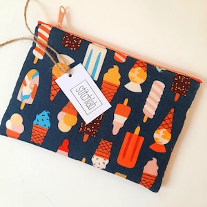 May include: A rectangular fabric zipper pouch featuring a dark blue background with a repeating pattern of colorful ice cream cones and popsicles. An orange zipper runs along the top edge. A white rectangular tag with the text stitchlab is attached.