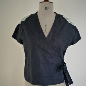 May include: A dark charcoal gray wrap top with short sleeves and a V-neckline. The garment features a textured fabric finish and contrasting light green and white floral patterned fabric panels on the shoulders. It ties at the side with a fabric belt.