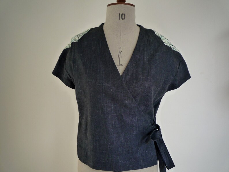 May include: A dark charcoal gray wrap top with short sleeves and a V-neckline. The garment features a textured fabric finish and contrasting light green and white floral patterned fabric panels on the shoulders. It ties at the side with a fabric belt.
